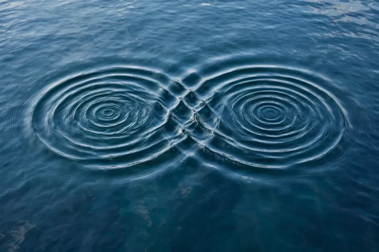 Two sets of circular ripples spreading across blue water overlap and interfere with each other, showing the superposition of waves where the ripples cross.